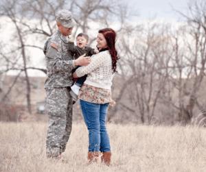 a military man and his wife in a field and holding their baby