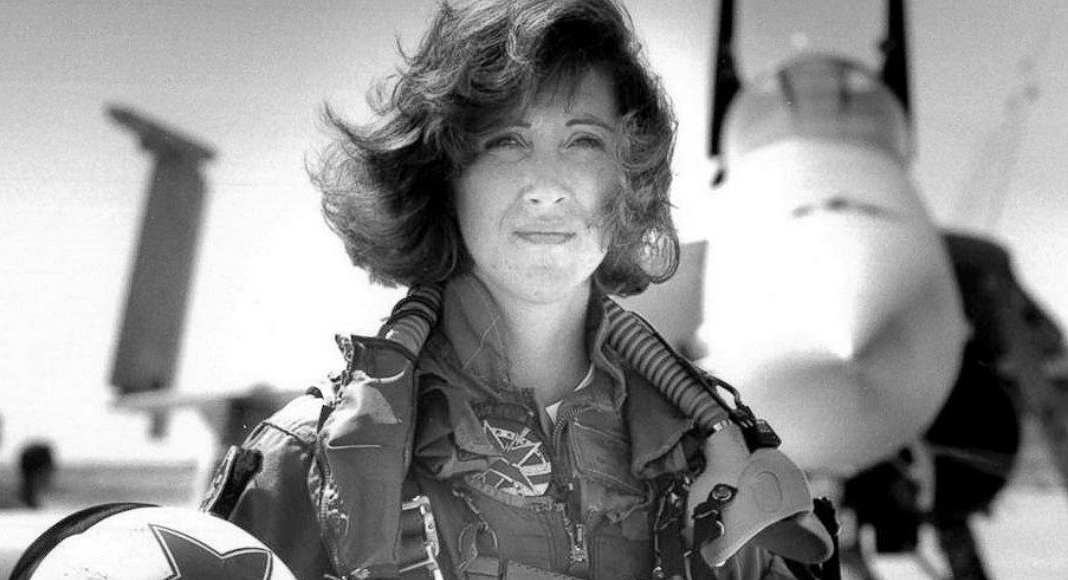 Female fighter pilot standing outside jet in uniform while holding helmet under arm