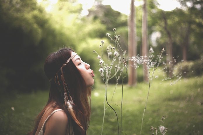 dark-haired woman blowing dandelion seeds in a field