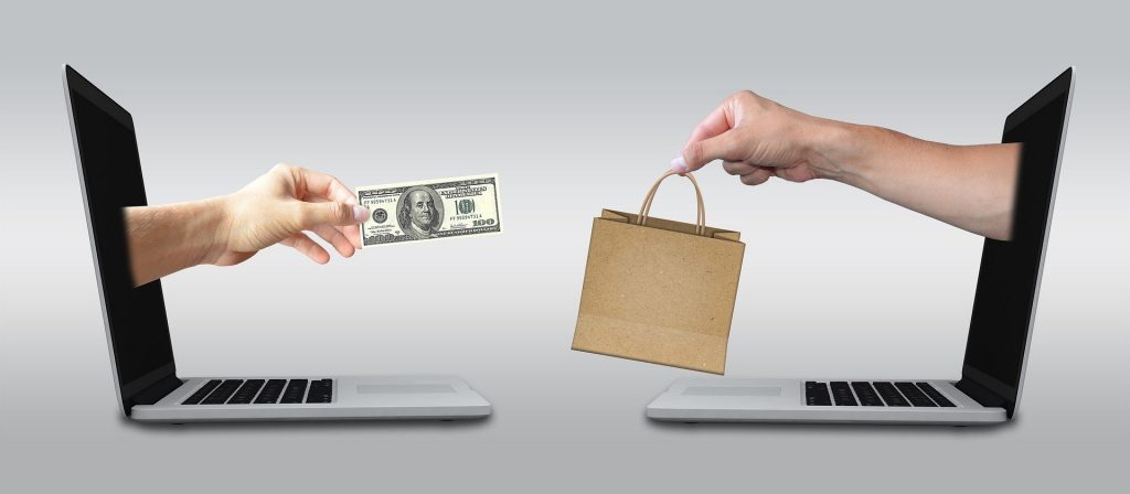 two laptops with hands reaching out, one handing cash to another handing a shopping bag