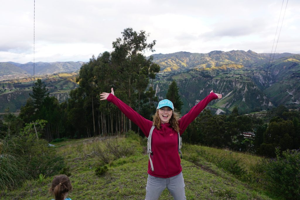 blonde woman in a red jacket and blue baseball cap standing in front of mountains