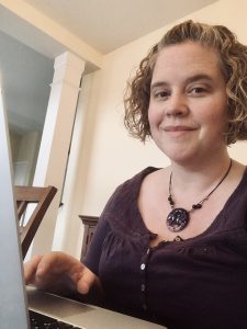 A white woman with short, curly hair sits at a laptop writing.