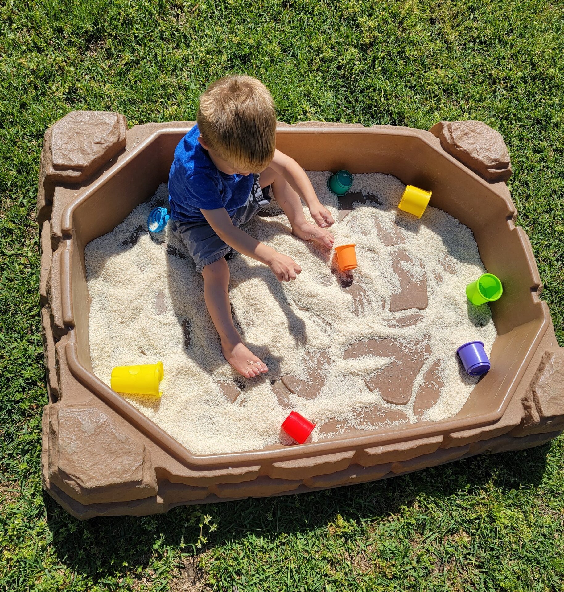 Little boy sitting in sand box playing with rice and colorful cups