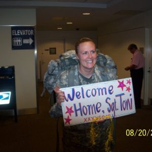 female service member in unifrom coming home from Iraq, smiling and holding a welcome home sign