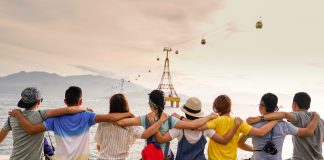 8 young people: 4 women and 4 men, sit in a line with their arms around the shoulders of the person next to them. Their backs are facing the camera as they look into the distance. The sky is bright and slightly orange.