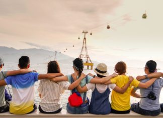 8 young people: 4 women and 4 men, sit in a line with their arms around the shoulders of the person next to them. Their backs are facing the camera as they look into the distance. The sky is bright and slightly orange.