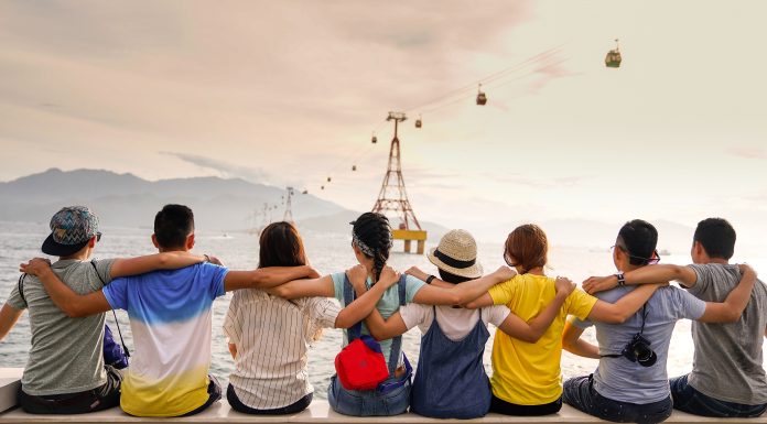 8 young people: 4 women and 4 men, sit in a line with their arms around the shoulders of the person next to them. Their backs are facing the camera as they look into the distance. The sky is bright and slightly orange.