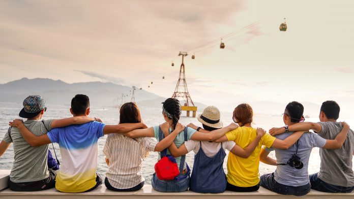 8 young people: 4 women and 4 men, sit in a line with their arms around the shoulders of the person next to them. Their backs are facing the camera as they look into the distance. The sky is bright and slightly orange.