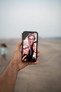 A hand and forearm can be seen holding a mobile phone. There is a video call in progress with three people on the screen. They are all smiling.