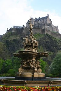 An ornate fountain is in the foreground surrounded by flowers. Edinburgh castle, Scotland, stands on a hill in the background.