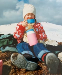 A girl sits on grass near a snowy mountain top in Scotland. She is wearing jeans and a red/white jumper and a white knitted hat. She is drinking from a pink and blue flask.