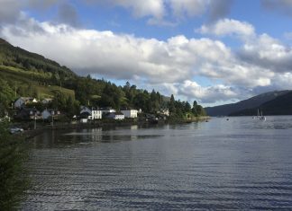 A coastal village in scotland sits on the banks of a loch. There are two sailing boats in the distance.