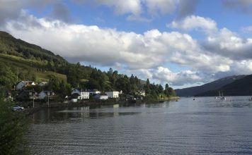 Friday Favorites – Top 5 Places to Visit in Scotland A coastal village in scotland sits on the banks of a loch. There are two sailing boats in the distance.