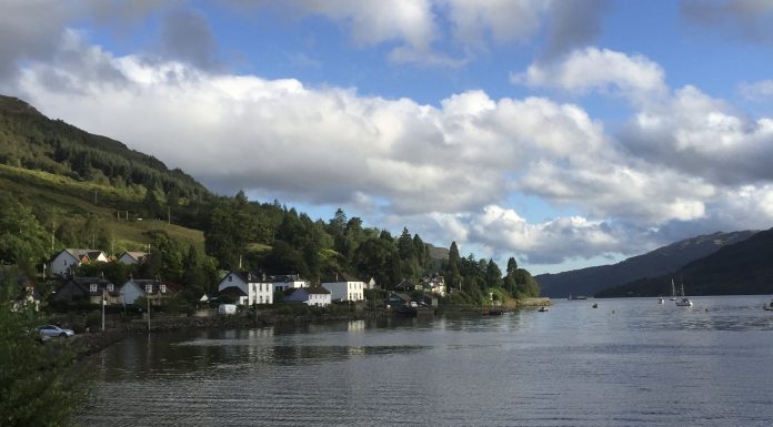 A coastal village in scotland sits on the banks of a loch. There are two sailing boats in the distance.