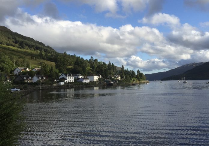 A coastal village in scotland sits on the banks of a loch. There are two sailing boats in the distance.