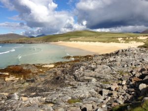 A rocky shore is in the foreground. In the background is a sandy beach and clear, blue water. The sky is bright blue with white clouds.