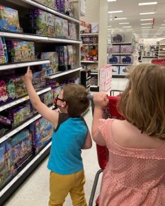 A girl in a peach dress is in the foreground with her back to the camera, she is pushing a red cart. There is a younger boy in front of her, also with his back to the camera, pointing and lego sets on a shelf.