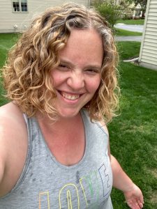 A caucasian woman with blonde, curly hair is wearing a grey vest. Her top has splashes of water on it and she has a wide smile on her face.