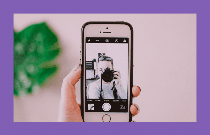 woman taking a selfie with camera and phone on a pink background and green plant shadowed behind it