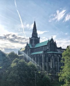 A large, dark, gothic style building sits amongst green trees. It has a green room and a spire reaches into the blue sky above.