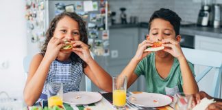 two children sitting at a table and eating sandwiches