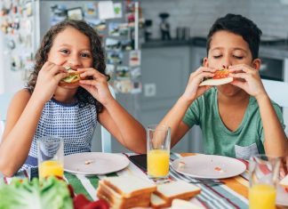 two children sitting at a table and eating sandwiches