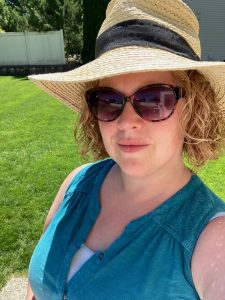 A white woman with curly hair stands in the sun. She is wearing sunglasses and a wicker hat.