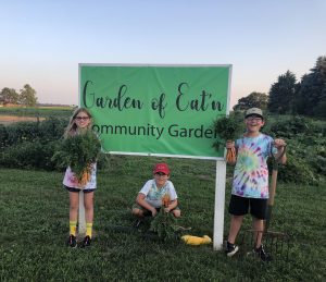 three children stand in front of a green sign saying Garden of Eat'n while holding bunches of carrots