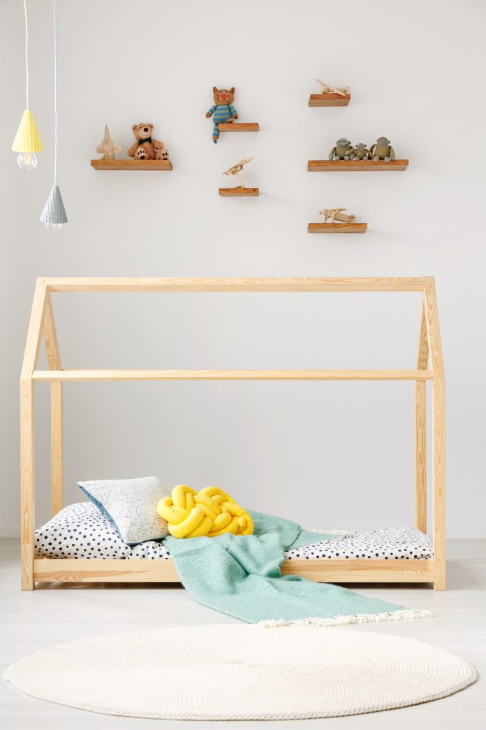 Natural wood Montessori-style toddler bed with pillows, small blanket. Toys sit on shelves mounted high above the bed.