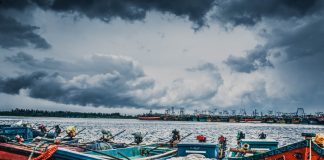 Wooden boats can be seen in the foreground with storm clouds in the sky behind them.