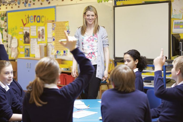 teacher standing in a classroom with students raising their hands