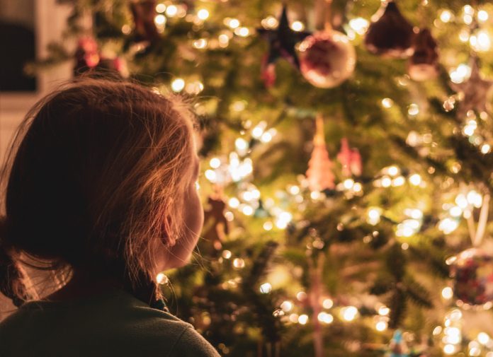 A little girl stands in the foreground looking away from camera towards a well-lit christmas tree.