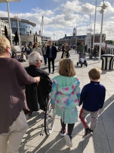 A lady is pushing a man in a wheelchair, walking away from the camera. Beside them their grandchildren are walking with them.