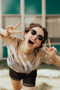 A woman in sunglasses is smiling and both of her hands are making peace signs.