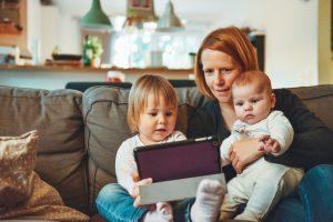 A woman sits with a toddler and a baby on her lap, looking at a laptop screen.