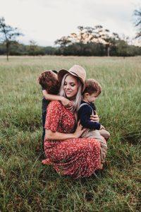 A woman is cuddling one child in front of her and another is cuddling her from behind. She is smiling.