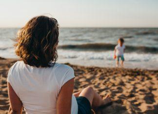 A mother sits in the foreground facing away from the camera. She is on a sandy beach and is watching her child paddling in the water, in the distance.