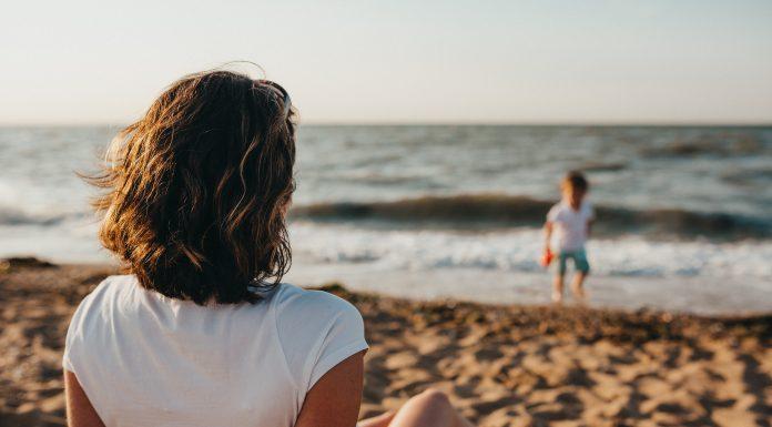 A mother sits in the foreground facing away from the camera. She is on a sandy beach and is watching her child paddling in the water, in the distance.