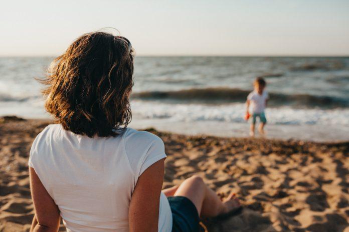 A mother sits in the foreground facing away from the camera. She is on a sandy beach and is watching her child paddling in the water, in the distance.