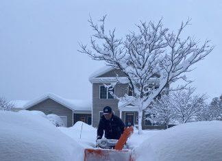 Man using snow blower to clear walk way in several feet of snow.