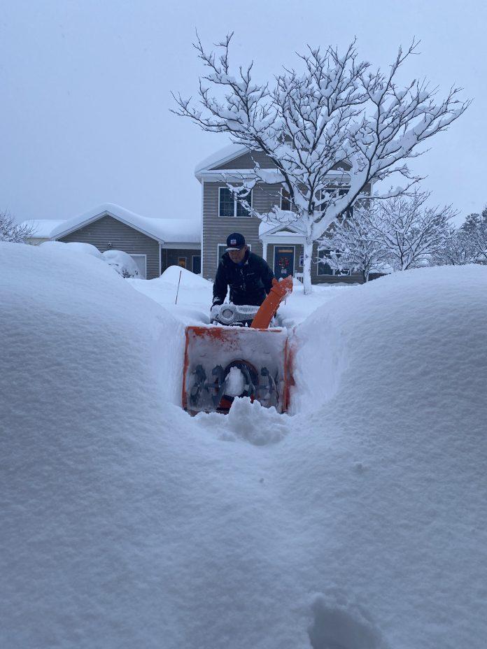 Man using snow blower to clear walk way in several feet of snow.