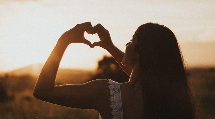 The silhouette of a woman can be seen against the setting sun. She is holding up her hands, forming a heart shape with her fingers.