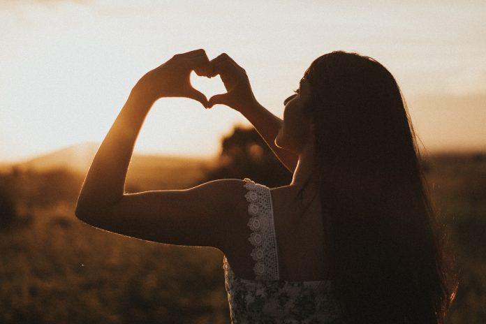 The silhouette of a woman can be seen against the setting sun. She is holding up her hands, forming a heart shape with her fingers.