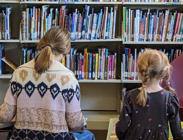 Two children read books in front of library shelves.