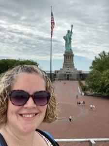 A white woman with curly hair, wearing sunglasses, is in the foreground. In the background is the American flag and the Statue of Liberty.