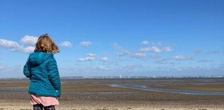 A young girl stands in the foreground, facing away from camera. She is looking out over a deserted beach on a clear day.