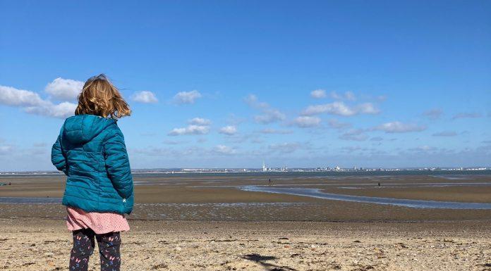 A young girl stands in the foreground, facing away from camera. She is looking out over a deserted beach on a clear day.