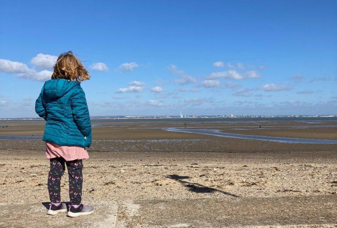 A young girl stands in the foreground, facing away from camera. She is looking out over a deserted beach on a clear day.