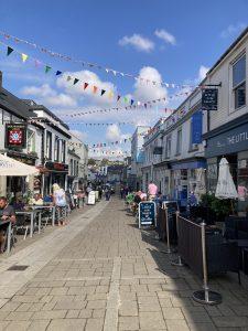 A village street is seen with shops on either side. There is colourful bunting over the street.