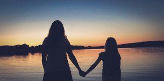 A silouette of a woman and young girl are seen, facing away from camera towards a sunset in the distance. They are holding hands.